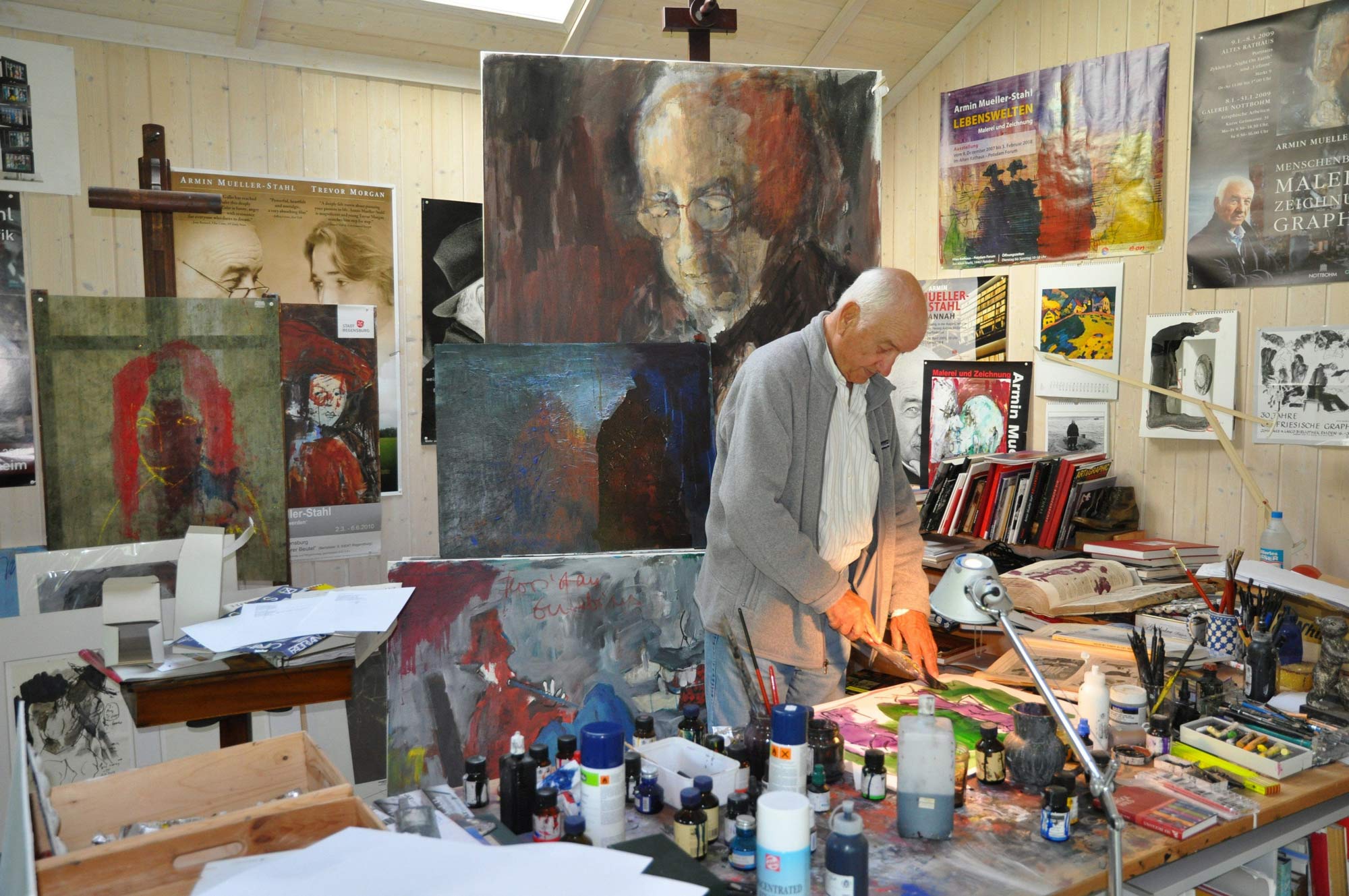 Armin Mueller-Stahl steht mit seinem Pinsel in der Hand in seinem Atelier, viele bunte Werke im Hintergrund